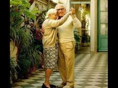 an older man and woman are hugging in front of some potted plants on the porch