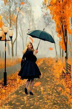 a woman in a black dress holding an umbrella while walking down a path with fall leaves on the ground