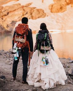 two people with backpacks walking down a path by the water in their wedding dresses