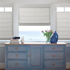 a blue dresser with books and vases on it in front of a large window