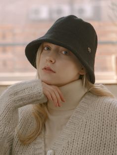 a woman wearing a hat and sweater looks off into the distance with her hand on her chin