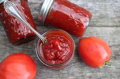 three tomatoes and two jars of tomato sauce on a wooden table with spoons in them