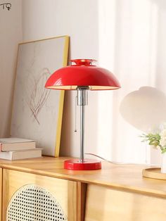 a red table lamp sitting on top of a wooden dresser next to a white vase