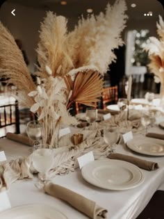 the table is set with white plates and silverware, tall pamodia stalks in vases