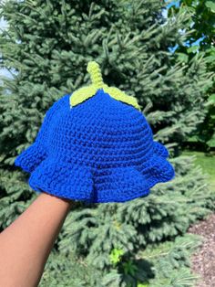 a crocheted blue hat is held up by someone's hand in front of a tree