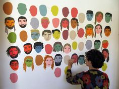 a woman standing in front of a wall with many different faces on it and holding her hand up to the wall