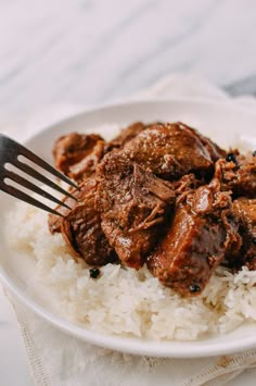 a white plate topped with rice and meat on top of a wooden table next to a fork