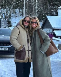 two women standing next to each other in front of snow covered ground and trees with cars behind them