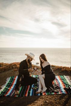 a man and woman sitting on top of a blanket next to the ocean