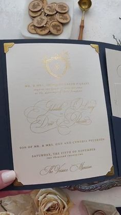 a person holding up a wedding card in front of some flowers and other things on the table