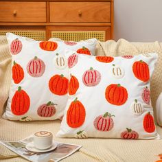 two pillows with pumpkins on them sitting on a bed next to a cup of coffee