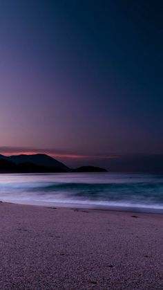 the sun is setting over the ocean with mountains in the distance and waves crashing on the beach