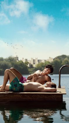 two people are laying on a dock in the water and one person is wearing swim trunks