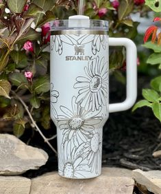 a stainless steel travel mug sitting on top of a stone wall next to some flowers