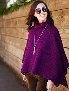 a woman standing next to a wooden fence wearing a purple ponchle and brown pants