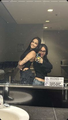 two women taking a selfie in a bathroom mirror with a sign on the wall