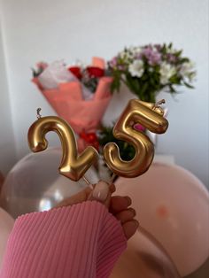 a person holding up two gold balloons with the number twenty five on them in front of a bouquet of flowers