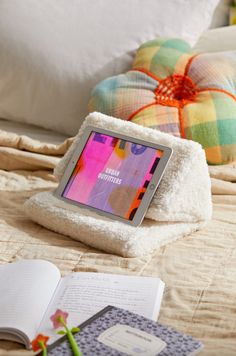 a tablet sitting on top of a bed next to an open book and flower vase