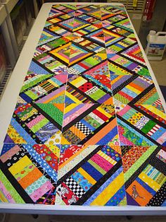 a table with a very colorful quilt on it's top and some shelves in the background