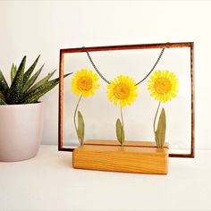 three yellow flowers are placed in front of a wooden frame with a plant on the side