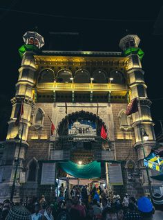 people are standing in front of a large building at night with lights on and flags hanging from it