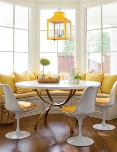 a white table with yellow cushions and chairs around it in front of two large windows