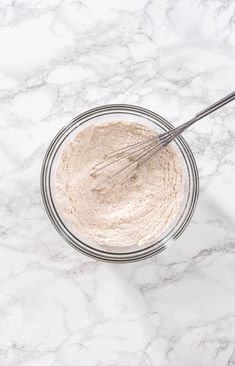 a glass bowl filled with dry ingredients on top of a white marble countertop next to a whisk