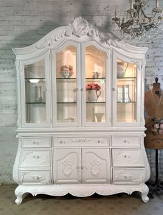 a white china cabinet with glass doors and flowers on the top, next to a mannequin