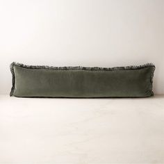 a long rectangular pillow with fringe trim on the bottom and sides, sitting against a white wall