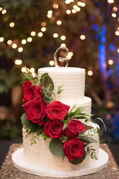 a white wedding cake with red roses on top and greenery around the edges, surrounded by lights