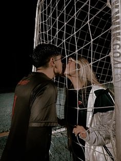 a man and woman kissing in front of a soccer ball net at night with the lights on