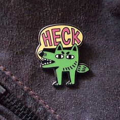 a green and black enamel pin with the word heck on it