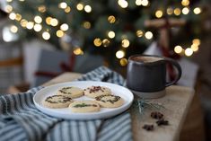 cookies on a plate next to a cup of coffee and a christmas tree with lights in the background