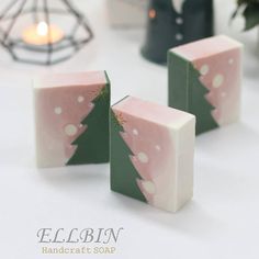 three pink and green christmas themed soaps on a white surface with a candle in the background