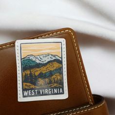 the west virginia sticker is on top of a brown leather bag with a white t - shirt in the background