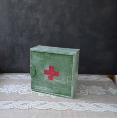 an old green box with a red cross painted on the side sitting on a white lace tablecloth