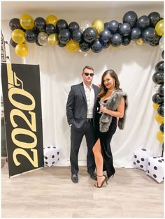 two people standing next to each other in front of a backdrop with black and gold balloons