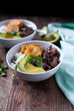 two bowls filled with meat and vegetables on top of a wooden table next to an avocado