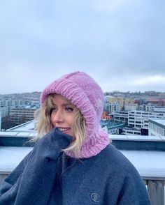 a woman with blonde hair wearing a pink knitted hat and blue sweater on top of a building