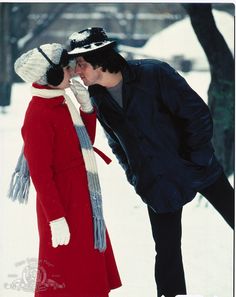 a man kissing a woman in the snow wearing a red coat and white hat with her hand on her face