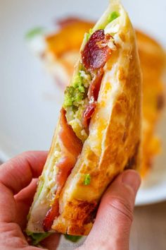 a hand holding a half eaten sandwich with broccoli and bacon on it in front of a plate