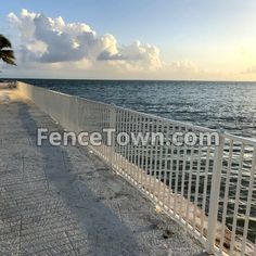 a white fence next to the ocean with clouds in the sky and palm trees on either side