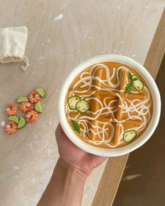 a person holding a bowl of soup with noodles and cucumbers in it on a table