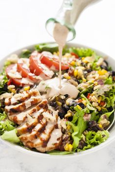 a salad with dressing being drizzled over it and topped with chicken, lettuce, tomatoes, black beans, corn, tomato, and more