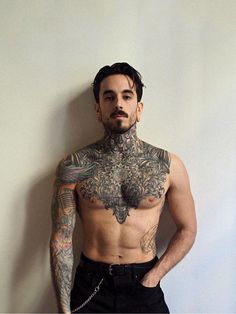 a man with tattoos on his chest and arms standing next to a wall wearing black pants