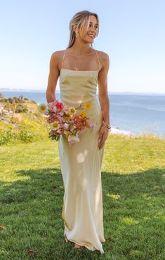 a woman in a long dress holding a bouquet of flowers standing on grass near the ocean