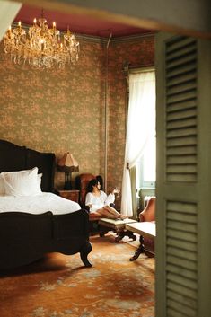 a woman sitting on a bed in a room with a chandelier hanging from the ceiling