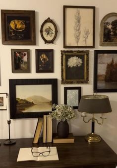 there are many framed pictures on the wall next to a table with a lamp and books