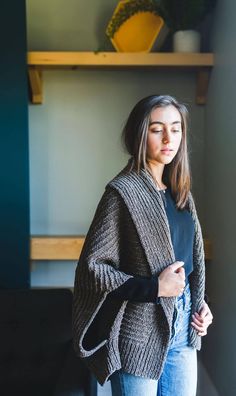 a woman standing in front of a shelf wearing a gray cardigan sweater and jeans