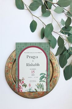 a wedding card on a gold plate next to some green leaves and branches in front of a white wall